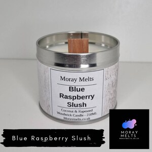 May include: A silver candle tin with a wooden lid. The label reads "Moray Melts Blue Raspberry Slush Coconut & Rapeseed Woodwick Candle - 250ML Moraymelts.co.uk".