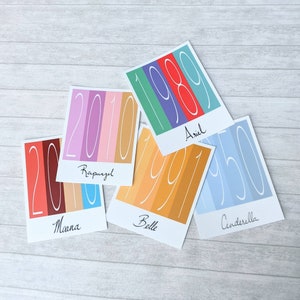 May include: Five colorful cards with dates and names. Each card features a different color scheme and a large year, such as 1989, 1950, 1991, 2010, and the names Ariel, Cinderella, Belle, and Rapunzel.