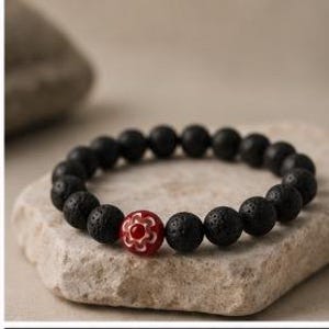 May include: A stretch bracelet composed of round, porous black lava stone beads. A single focal bead is included, featuring a deep red center surrounded by a white, flower-like petal pattern. The beads are strung together in a circular arrangement.