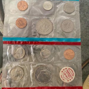 May include: A vintage uncirculated mint set of U.S. coins, sealed in clear plastic. The set includes various denominations, such as pennies, nickels, dimes, quarters, and half dollars. The packaging has red and blue stripes and a "Bureau of the Mint" sticker.