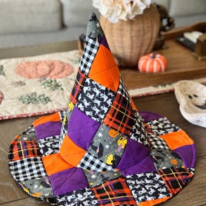 May include: A quilted witch hat featuring a patchwork design of orange, purple, and black and white plaid fabric squares. The fabric includes patterns of white ghosts, orange pumpkins, and black witch hats on a gray background with stitched details.