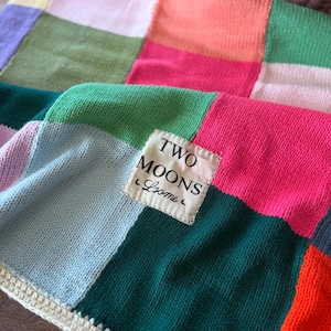 May include: A colorful patchwork blanket with squares of pink, green, blue, orange, and purple. The blanket has a cream-colored crocheted border and a label that reads "TWO MOONS Looms."