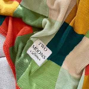 May include: A colorful patchwork blanket with a variety of colors including orange, yellow, green, and beige. The blanket has a label that reads "TWO MOONS Looms". The blanket is made of a knitted material.