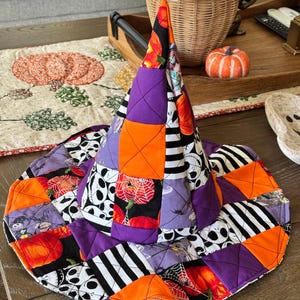 May include: A quilted witch hat featuring a patchwork design of orange, purple, and black fabric squares. The pattern includes illustrations of pumpkins, spider webs, and skull-like faces, with black and white stripes throughout the fabric.