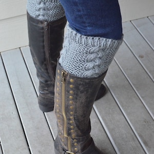 May include: Gray knitted boot cuffs with a cable knit pattern worn over black leather boots with a zipper and buckle.