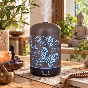 May include: A cylindrical dark brown essential oil diffuser with a cut-out butterfly and floral pattern that glows with a blue light. The device emits a steady stream of mist from the top and features the text Shay MG above control buttons.