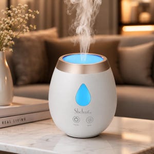 May include: A white and gold essential oil diffuser with a blue light. The word "Shehan" is printed on the front. The diffuser is on a white table. The diffuser emits steam. The table is in front of a sofa.