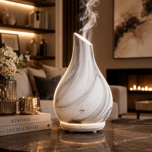 May include: A white and grey marble-effect essential oil diffuser with a distinctive angled top. The diffuser is emitting steam. The base is white with a soft, glowing light. The product is on a dark marble surface.