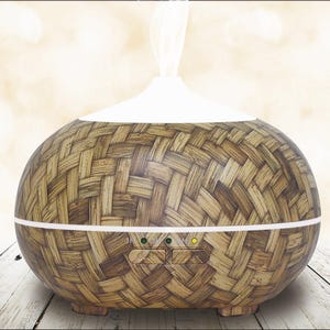 May include: An oval essential oil diffuser with a woven wood-effect finish. The diffuser has a white top and a white band around the middle. Two green and yellow lights are visible on the front. A wisp of vapor rises from the top.