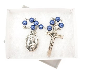 Op de afbeelding: Een zilveren rozenkrans met blauwe parels in een witte doos. De rozenkrans heeft een zilveren kruisbeeld en een zilveren medaille van de Maagd Maria.