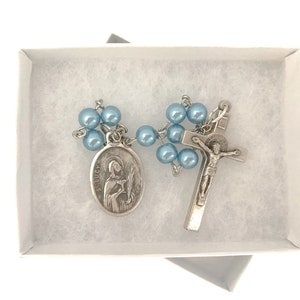 May include: A light blue rosary with a silver crucifix and a silver medal of Saint Lucy. The rosary is in a white box with white tissue paper.