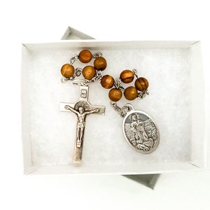 May include: A wooden rosary with brown beads, a silver crucifix, and a silver medal with a religious image. The rosary is in a white box with white tissue paper.
