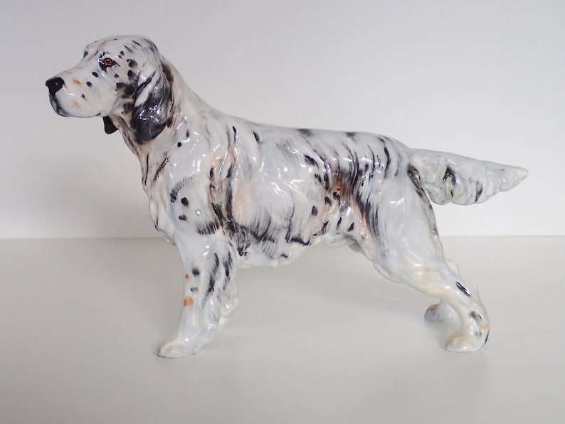May include: A white and black porcelain figurine of a hunting dog, a setter, standing with its head turned to the left. The dog has brown eyes and a black nose.