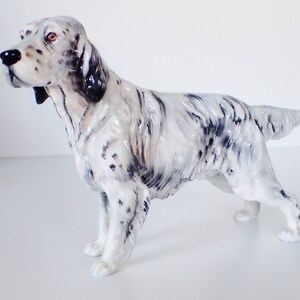 May include: A white and black ceramic figurine of a hunting dog, a pointer, standing with its head turned to the left. The dog has a black nose and black markings on its face and body.
