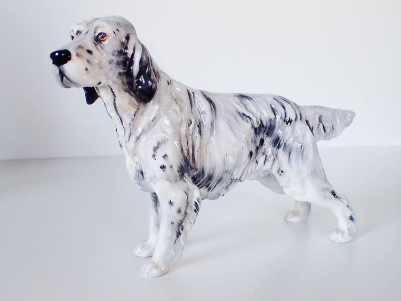 May include: A white and black ceramic figurine of a hunting dog, a pointer, standing with its head turned to the left. The dog has a black nose and black markings on its face and body.