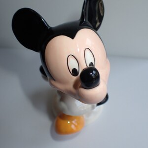 Op de afbeelding: Een keramiek beeldje van Mickey Mouse. Het beeldje is geschilderd in zwart, wit en geel. Mickey Mouse draagt zijn kenmerkende rode broek en gele schoenen. Hij heeft een grote glimlach op zijn gezicht.
