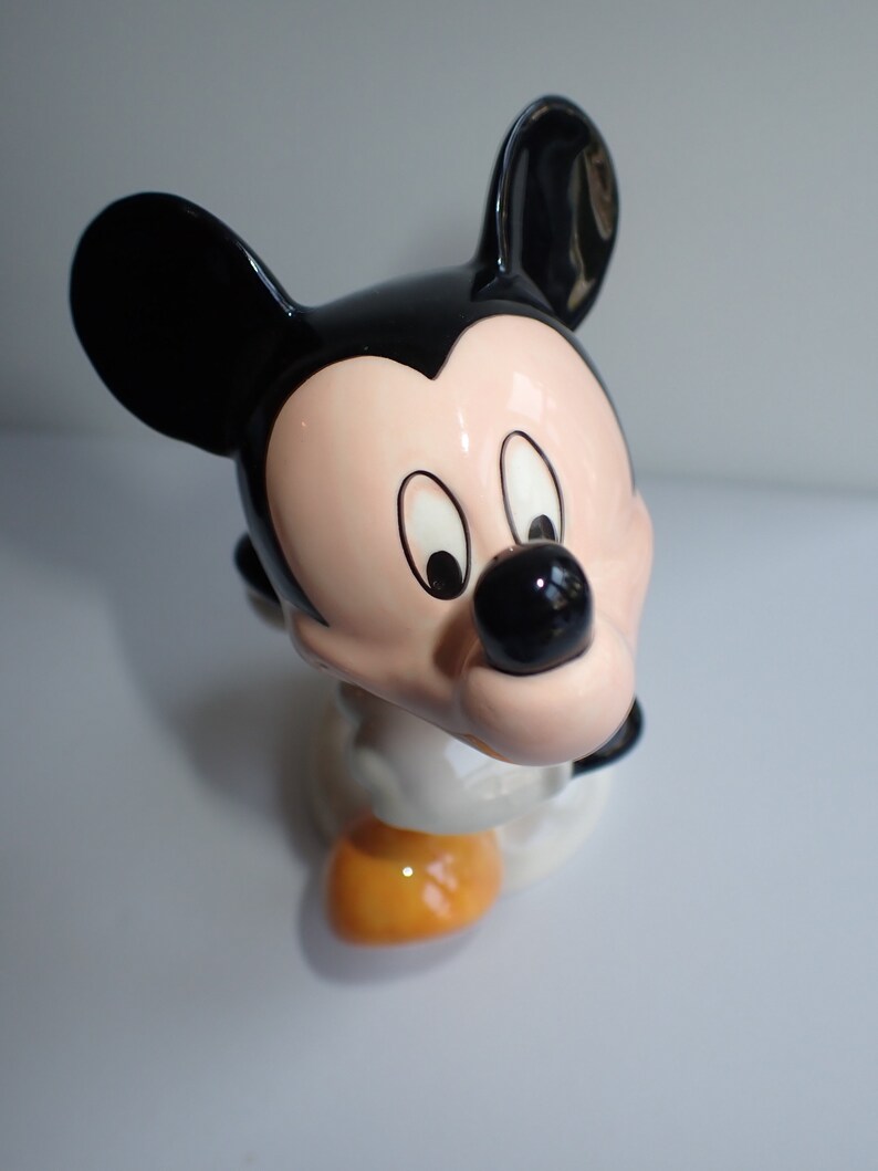 Op de afbeelding: Een keramiek beeldje van Mickey Mouse. Het beeldje is geschilderd in zwart, wit en geel. Mickey Mouse draagt zijn kenmerkende rode broek en gele schoenen. Hij heeft een grote glimlach op zijn gezicht.