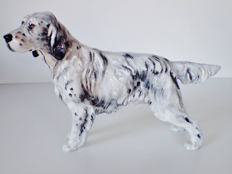 May include: A white and black porcelain figurine of a standing English Setter dog. The dog is posed with its head turned to the left and its tail curled up.