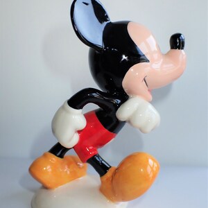 Op de afbeelding: Een keramiek beeldje van Mickey Mouse in een looppose. Hij draagt zijn kenmerkende rode broek, witte handschoenen en gele schoenen. Zijn oren zijn zwart en zijn gezicht is lichtroze.