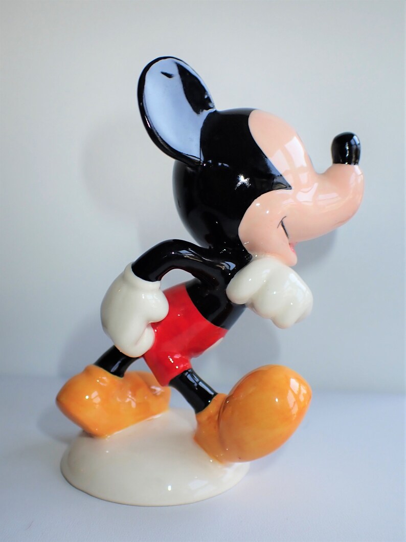 Op de afbeelding: Een keramiek beeldje van Mickey Mouse in een looppose. Hij draagt zijn kenmerkende rode broek, witte handschoenen en gele schoenen. Zijn oren zijn zwart en zijn gezicht is lichtroze.