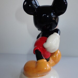 Op de afbeelding: Een keramiek beeldje van Mickey Mouse dat op een witte basis staat. Mickey draagt zijn kenmerkende rode broek, gele schoenen en witte handschoenen. Hij heeft zwarte oren en een zwart gezicht.
