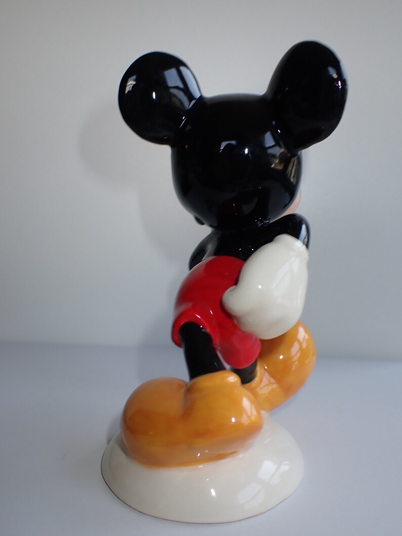 Op de afbeelding: Een keramiek beeldje van Mickey Mouse dat op een witte basis staat. Mickey draagt zijn kenmerkende rode broek, gele schoenen en witte handschoenen. Hij heeft zwarte oren en een zwart gezicht.