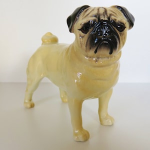 May include: A ceramic figurine of a pug dog. The pug is standing with its front legs slightly apart and its back legs together. The pug is light brown with black ears and a black face. The figurine is detailed with wrinkles on the face and a curled tail.
