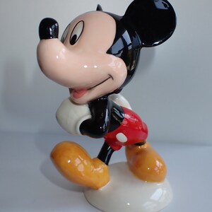 Op de afbeelding: Een keramiek beeldje van Mickey Mouse, staand met zijn rechterbeen naar voren en zijn linkerarm gebogen. Hij draagt een rode broek met witte stippen, gele schoenen en een witte handschoen aan zijn linkerhand. Zijn gezicht is geschilderd met zwarte ogen, een rode neus en een brede glimlach.