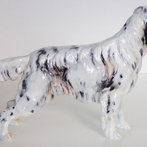 May include: A white and black ceramic figurine of a dog, possibly a setter, standing on a white surface. The figurine is detailed with black markings and a glossy finish.