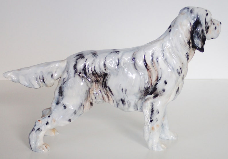 May include: A white and black ceramic figurine of a dog, possibly a setter, standing on a white surface. The figurine is detailed with black markings and a glossy finish.