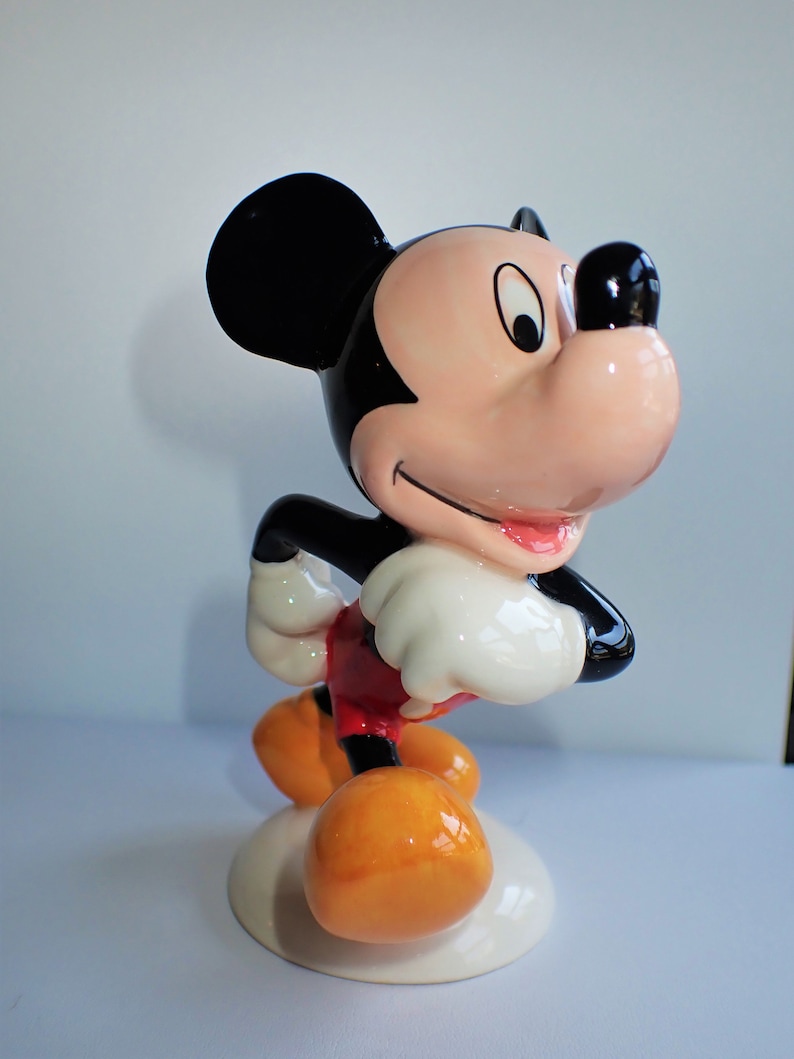 Op de afbeelding: Een keramiek beeldje van Mickey Mouse, staand met gekruiste armen en een glimlach op zijn gezicht. Hij draagt zijn kenmerkende rode broek en gele schoenen.