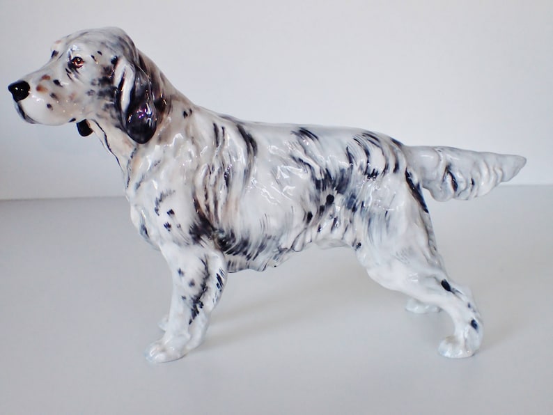 May include: A white and black porcelain figurine of a standing English Setter dog. The dog is posed with its head turned to the left and its tail curled.