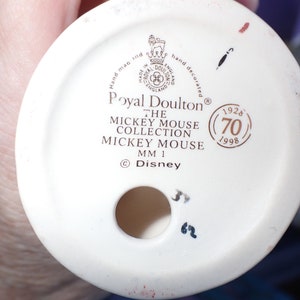 Op de afbeelding: Een witte keramische figuurbasis met gouden letters die "Royal Doulton&reg; The Mickey Mouse Collection Mickey Mouse MM 1 &copy;Disney" luidt en een gouden cirkel met de tekst "1928 (70) 1998".