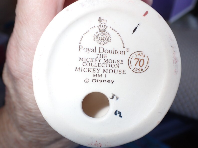 Op de afbeelding: Een witte keramische figuurbasis met gouden letters die "Royal Doulton&reg; The Mickey Mouse Collection Mickey Mouse MM 1 &copy;Disney" luidt en een gouden cirkel met de tekst "1928 (70) 1998".