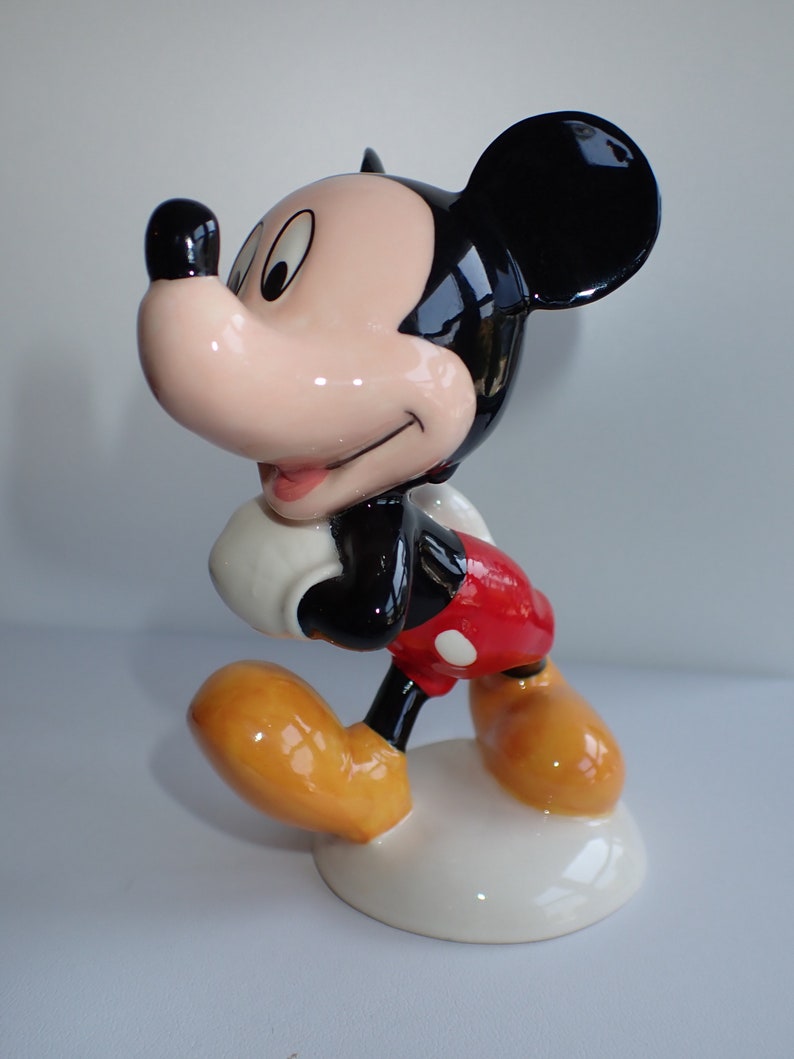 Op de afbeelding: Een keramiek beeldje van Mickey Mouse in een looppose. Hij draagt zijn kenmerkende rode broek met witte stippen, gele schoenen en witte handschoenen. Zijn oren zijn zwart en zijn gezicht is lichtroze.