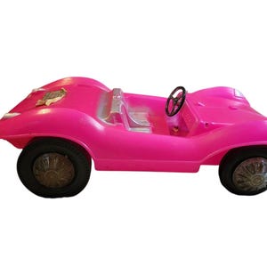 Puede incluir: Un coche de juguete vintage de color rosa brillante con neumáticos negros y un volante negro. El coche tiene un diseño de dos plazas y una estética clásica. Las ruedas tienen un diseño de tapacubos plateado. El coche es de plástico.