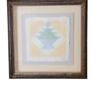 May include: A framed artwork featuring a hand-stitched quilt block with a geometric design in shades of blue, green, and yellow. The quilt block is surrounded by a white border and a light gray mat.