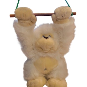 May include: A plush white monkey toy with a light brown wooden bar and green and blue rope. The monkey has a smiling face and is holding onto the bar with its hands. The toy is designed to hang.