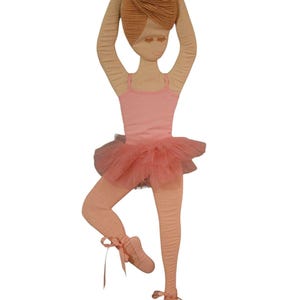May include: A fabric ballerina doll in a graceful pose. The doll wears a pink leotard, a tulle tutu, and ballet slippers with ribbons. Her hair is styled in a bun, and her arms are raised above her head.