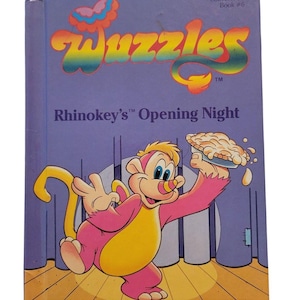 Könnte beinhalten: Ein Vintage-Buch "Wuzzles" mit dem Titel "Rhinokey's Opening Night". Das Cover ist lila mit farbenfrohem Text und einer Illustration eines rosa-gelben Cartoon-Charakters, der einen Kuchen hält. "Collector Series Book #6" ist oben aufgedruckt.