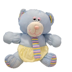 May include: A light blue plush teddy bear with a white muzzle and pink nose. The bear wears a yellow diaper and a striped tie with pink, blue, and yellow stripes. The bear's ears and feet have striped accents.