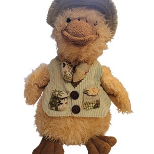 May include: A plush duck toy wearing a green corduroy vest with two pockets and brown buttons. The duck has a matching hat and a patterned bow tie. The duck's body is a light brown color, and it has brown feet.