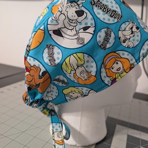 May include: A blue surgical scrub cap featuring cartoon characters from Scooby-Doo. The cap has a repeating pattern of Scooby-Doo, Fred, Daphne, and the words "Scooby-Doo!" in white circles. The cap is tied at the back.