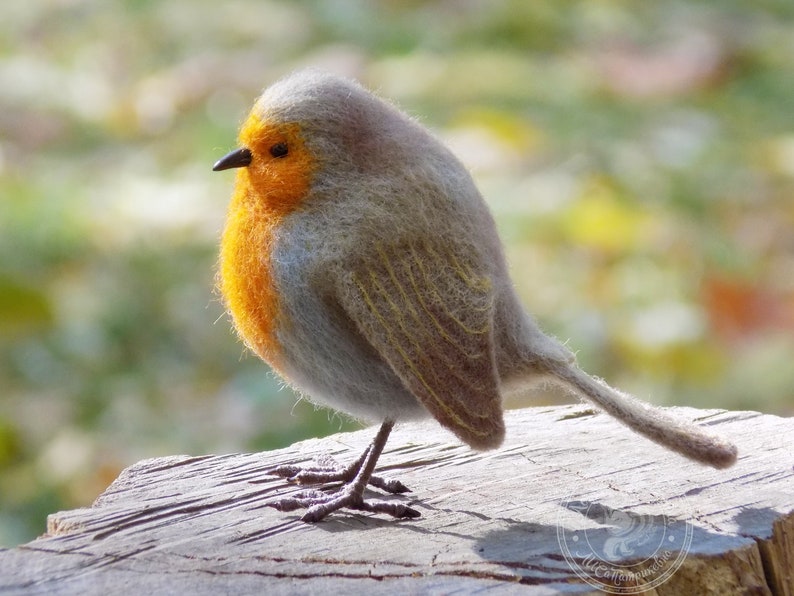 May include: A small, gray and orange robin bird figurine made of felt. The bird is perched on a wooden surface.