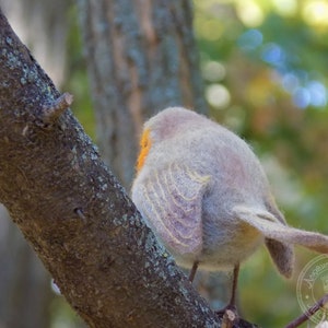 May include: A gray and white felted bird perched on a tree branch. The bird has a yellow beak and orange markings on its chest. The bird is facing away from the camera.