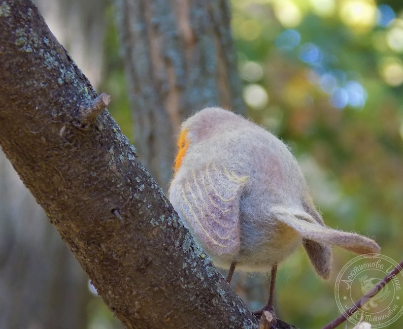 May include: A gray and white felted bird perched on a tree branch. The bird has a yellow beak and orange markings on its chest. The bird is facing away from the camera.