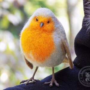 May include: A close-up of a needle felted robin, a small bird with a gray head and a bright orange breast. The robin is perched on a dark surface.