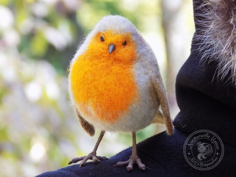 May include: A close-up of a needle felted robin, a small bird with a gray head and a bright orange breast. The robin is perched on a dark surface.
