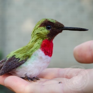 May include: A handmade hummingbird figurine with green, red, and white feathers. The bird has a long, black beak and is perched on a hand. The hummingbird's chest is a vibrant red, and its wings are a mix of green and brown.