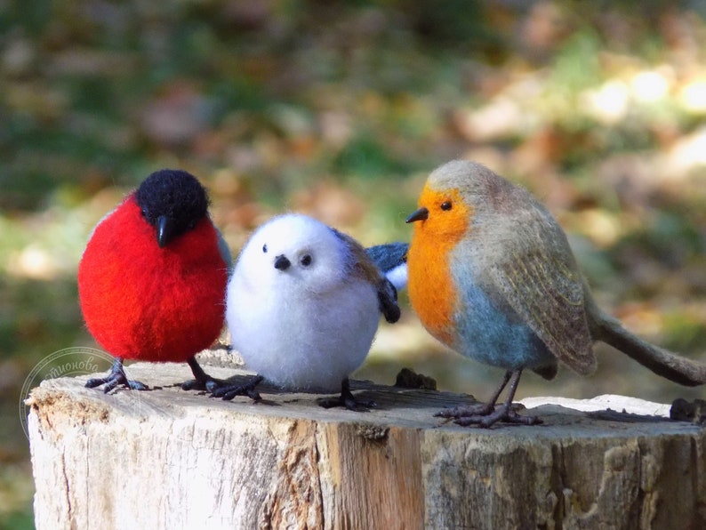 May include: Three felt bird figurines, a red robin, a white chickadee, and a blue robin, perched on a tree stump.